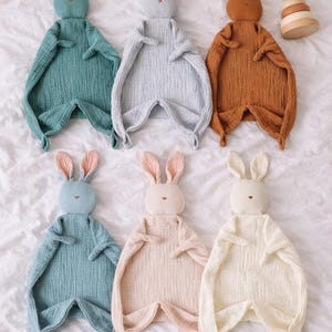 Könnte beinhalten: Eine Auswahl an Baby-Kuscheltüchern in Hasenform und Pastellfarben wie Blau, Grau und Rosa, auf einer weißen Oberfläche angeordnet. Jedes Kuscheltuch hat einen Hasenkopf mit langen Ohren und einen strukturierten Körper. Ein Holz-Steckspielzeug ist ebenfalls vorhanden.