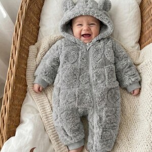 Peut inclure: Un bébé souriant portant une combinaison grise à capuche avec des oreilles d'ours, allongé dans un panier en osier. La combinaison a une fermeture éclair et des poignets côtelés. Le bébé est sur un oreiller blanc et une couverture crème. Le logo MiniMuse Atelier est visible.