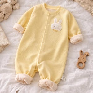 Peut inclure: Combinaison jaune pour bébé avec un appliqué lapin. Le vêtement une pièce à manches longues et côtelé a des poignets crème et une fermeture boutonnée. Un anneau de dentition en bois en forme de lapin et une peluche lapin sont également visibles. Le texte "MiniMuse ATELIER" est visible.