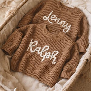 Può includere: Due maglioni marroni a maglia con nomi ricamati bianchi, "Lenny" e "Ralph", sono in un cestino intrecciato. I maglioni hanno polsini a coste e girocollo. Il cestino è foderato con tessuto color crema. Il logo MiniMuse Atelier è visibile.