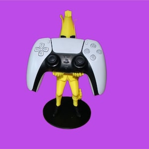 May include: A yellow banana-shaped figurine holding a white and black video game controller. The figurine stands on a black circular base against a purple background. The controller has a PlayStation logo.