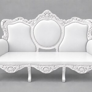 May include: A white sofa featuring an ornate, carved wooden frame with intricate floral and shell motifs along the backrest and base. The piece has a smooth, solid white upholstered seat and backrest, supported by four curved legs with detailed carvings.