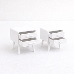 Puede incluir: Dos mesitas de noche blancas en miniatura, cada una con dos cajones. Las mesas tienen un diseño minimalista con bordes redondeados y patas cónicas. Una mesa tiene ambos cajones abiertos, la otra tiene uno abierto.