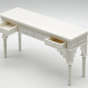 Op de afbeelding: Een witte miniatuurbureau met twee open lades. Het bureau heeft sierlijke details op de poten en ladefronten. Het bureau is rechthoekig en lijkt van een stevig materiaal te zijn gemaakt. De laden zijn leeg.