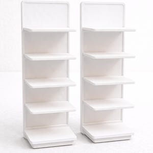 Op de afbeelding: Twee witte miniatuur displayrekken. Elk rek heeft een rechthoekige basis, vier planken en een rechthoekige bovenkant. De planken zijn gelijkmatig verdeeld en lijken gemaakt van een glad, stevig materiaal. Ideaal voor kleine items.