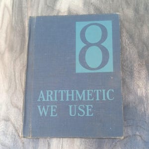 May include: A vintage textbook titled "Arithmetic We Use" with a large number 8 on the cover. The cover is a muted blue with the text and number in a contrasting teal color. The book appears to be well-worn.