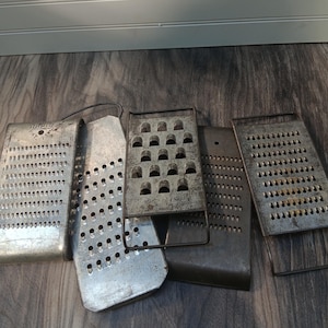 May include: A collection of vintage metal graters in various shapes and sizes. The graters are made of silver metal and have different hole patterns for grating food. Some graters have handles, and all show signs of wear.
