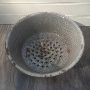 May include: A vintage, gray enamelware colander with a speckled pattern and a perforated base. The rim shows signs of wear, adding to its antique charm. This kitchenware item is perfect for draining pasta or washing vegetables.