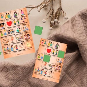 May include: Two orange cards with illustrations and Spanish text, featuring words like "amistad" and "corazón." A red heart is in the center of one card. Green squares and a brown fabric are in the background.