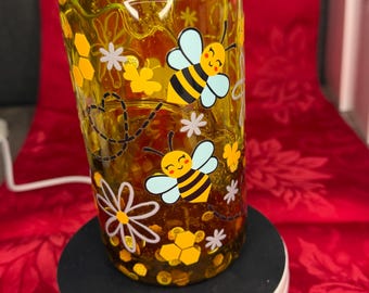 May include: A clear glass jar with a light green lid, decorated with yellow bees, flowers, and honeycomb patterns. The jar is filled with a yellow liquid and sits on a black and white rotating stand.