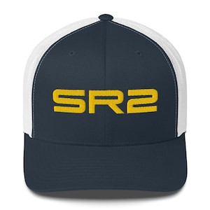 May include: Navy blue trucker hat with a white mesh back and a curved brim. The front of the hat features the embroidered text "SR2" in yellow. The hat has a structured front panel and an adjustable snapback closure.