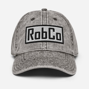 May include: A gray, vintage-style baseball cap with a curved brim. The cap features the embroidered text "RobCo" in black block letters inside a black rectangular frame. The hat has a slightly faded appearance.