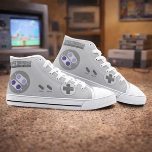 Könnte beinhalten: Hellgraue High-Top-Sneaker mit Super Controller-Design. Die Sneaker zeigen ein klassisches Videospiel-Controller-Layout mit Richtungspfeilen und Knöpfen in Lila und Grau. Weiße Schnürsenkel und Sohlen vervollständigen das Design.
