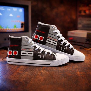 Könnte beinhalten: Paar High-Top-Sneaker mit Retro-Videospiel-Controller-Design. Die Sneaker haben ein Schwarz-Grau-Farbschema mit roten und weißen Akzenten, einschließlich des Wortes "Controller" und Spielknöpfen. Weiße Schnürsenkel und Sohlen vervollständigen den Look.