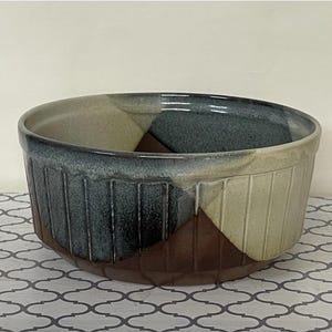 May include: A ceramic bowl with a geometric design. The bowl features a color-blocked pattern with sections of brown, beige, and teal. The exterior has vertical ribbed detailing. The interior is a light beige color.