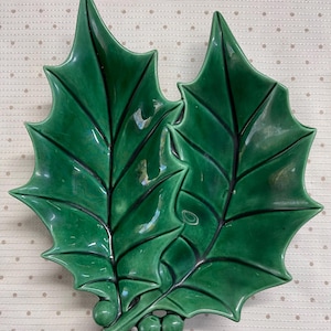 May include: A glossy green ceramic dish shaped like two holly leaves with three small berries at the base. The dish features dark green veins and a smooth, reflective surface, capturing the distinct pointed edges and organic form of holly foliage.