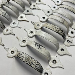 May include: A collection of arched metal drawer pulls arranged in rows. Each handle features a distressed white and black finish with a textured surface. The pulls have teardrop-shaped mounting plates at each end, each containing a single screw hole.