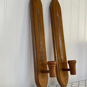 May include: A pair of matching wall-mounted candle holders made of medium-toned wood. Each holder features a long, pointed, shield-shaped backplate with a central vertical groove and a single cylindrical candle cup extending from the lower section.
