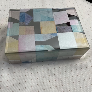 May include: A rectangular glass box with a lid. The lid is decorated with a patchwork design of various pastel-colored squares and rectangles. The box is sitting on a white surface with small gray polka dots.