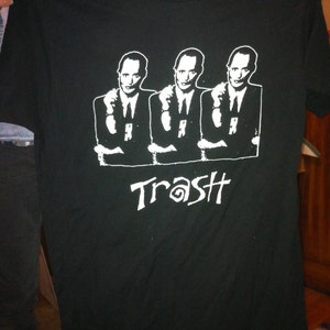 May include: Black t-shirt featuring a white graphic of three men in suits with the word "Trash" printed below.
