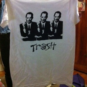 Op de afbeelding: Wit T-shirt met een zwart-witte grafische print van drie mannen in pak. De tekst "Trash" is onder de print gedrukt.