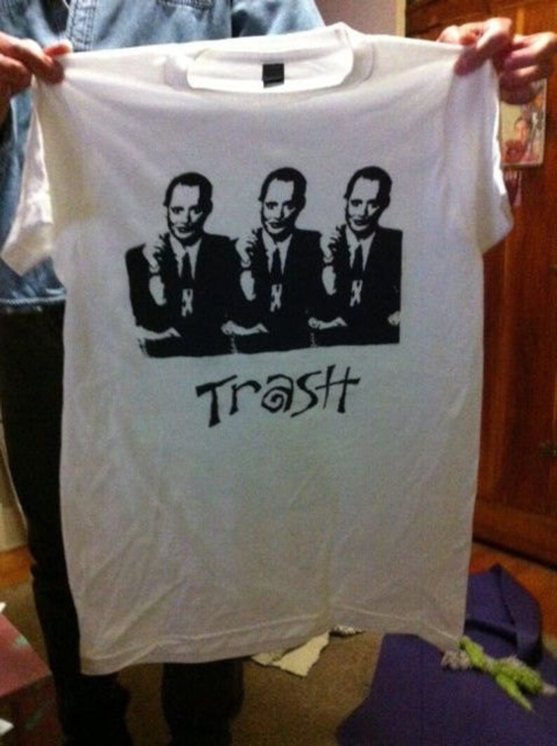 Op de afbeelding: Wit T-shirt met een zwart-witte grafische print van drie mannen in pak. De tekst "Trash" is onder de print gedrukt.