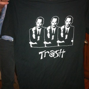 May include: Black t-shirt featuring a white graphic of three men in suits with their arms crossed. The text "Trash" is printed below the men.