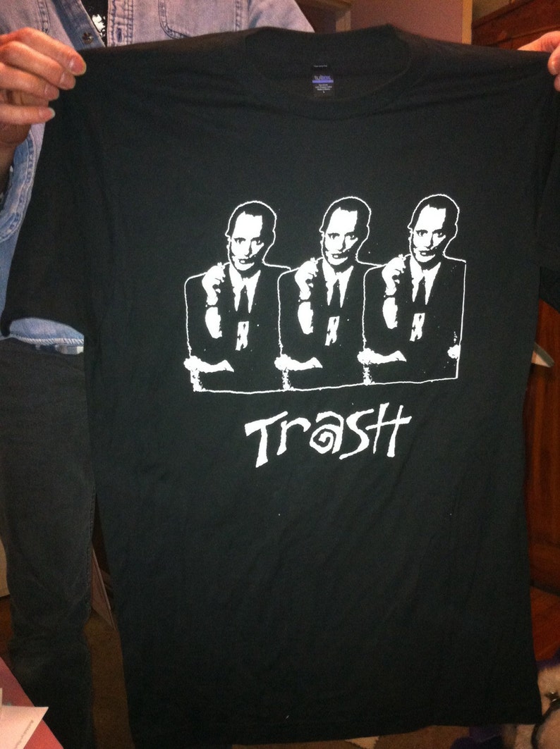 May include: Black t-shirt featuring a white graphic of three men in suits with their arms crossed. The text "Trash" is printed below the men.