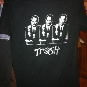 May include: Black t-shirt featuring a white graphic of three men in suits with their hands raised, pointing at the viewer. The text "Trash" is printed below the men in a stylised font.
