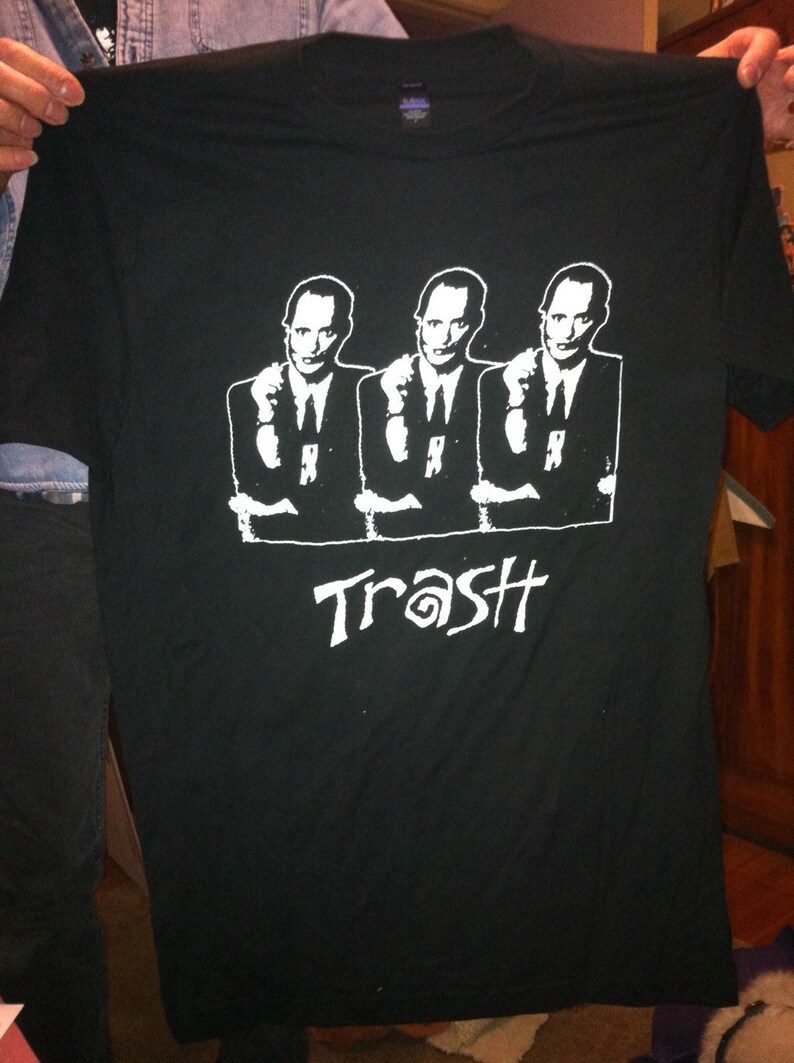 May include: Black t-shirt featuring a white graphic of three men in suits with their hands raised, pointing at the viewer. The text "Trash" is printed below the men in a stylised font.