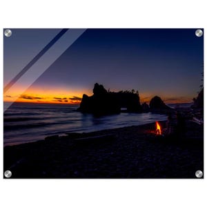 Könnte beinhalten: Ein Landschaftsfoto der Küste in der Abenddämmerung. Das Bild zeigt dunkle Silhouetten von Felsformationen und einen Strand mit einem kleinen Feuer. Der Himmel geht von Orange in tiefes Blau über und spiegelt sich im Wasser.