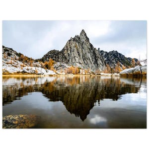 May include: A scenic landscape photograph featuring a mountain reflected in a calm lake. The mountain is gray and rocky, with a sharp peak. The water reflects the mountain and the cloudy sky. The scene includes trees with yellow leaves and patches of snow.