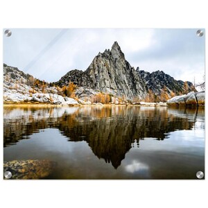 May include: A scenic landscape print featuring a mountain reflected in a calm lake. The mountain is gray and rocky, with a jagged peak. The water reflects the mountain and the cloudy sky. The image is mounted on a clear acrylic panel.