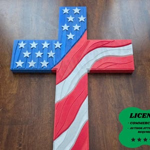 May include: A wooden cross with a patriotic design. The top and left arm of the cross are blue with white stars, while the rest features red and white stripes, resembling the American flag. The cross is placed on a wooden surface.