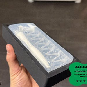 Puede incluir: Un objeto blanco en forma de tenedor está encerrado en un molde rectangular transparente dentro de una caja negra. La imagen incluye una pegatina verde con la palabra "LICENSE" y otro texto.