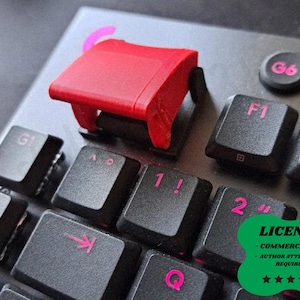 Op de afbeelding: Close-up van een zwart computertoetsenbord met roze letters en een felrode keycap. Het toetsenbord heeft toetsen met de labels G1, G2, F1, F2 en cijfers. Een groene sticker met het woord "LICENSE" staat rechtsonder.