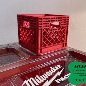 May include: A small, red plastic crate with a diamond pattern sits atop a red and clear container. The container has the Milwaukee Packout logo in white. The crate has two rectangular openings on the sides.