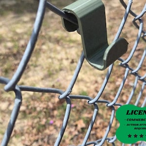 May include: A close-up of a green plastic fence hook attached to a chain-link fence. The hook is designed to hang items on the fence. The background is blurred, showing a grassy area. The image includes a green license badge.
