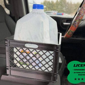 May include: A black plastic milk crate holding a large water jug with a blue cap, placed in a car. The crate has a diamond pattern and is positioned on a car seat. The steering wheel is visible in the background.