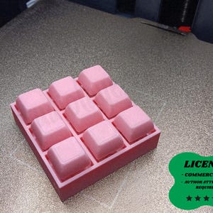 Puede incluir: Una bandeja rosa impresa en 3D con nueve compartimentos cuadrados elevados. La bandeja es de color rosa sólido y los compartimentos están espaciados uniformemente. La imagen también incluye una insignia de licencia verde con texto.