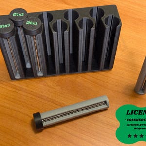 Puede incluir: Un organizador negro y gris impreso en 3D con compartimentos cilíndricos, que contiene varios objetos cilíndricos grises y negros. Los objetos tienen tapas negras con texto verde, incluyendo "Ø3x2", "Ø3x3" y "Ø6x3". Un objeto separado, etiquetado "Ø4x2".