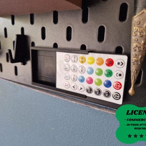 Può includere: Un telecomando in bianco e nero con pulsanti colorati è montato su una pannello forato nero. A destra c'è un oggetto decorativo in metallo con un design ornato. L'immagine include anche un badge di licenza verde.