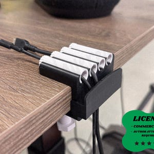 May include: A black desk cable organizer with white and black accents, holding several black charging cables. The organizer is clamped to a wooden desk. The image includes a green license badge.