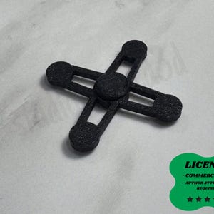 Puede incluir: Un objeto negro en forma de cruz con extremos redondeados y recortes rectangulares. El objeto está sobre una superficie blanca. Una pegatina verde con la palabra "LICENSE" y otro texto está en la esquina inferior derecha.