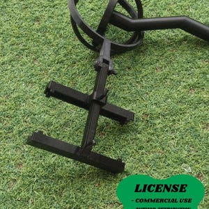 May include: A black metal object with a circular top and a cross-shaped base. The object has a handle and is set against a backdrop of green grass. The image includes a green oval with the word "LICENSE" and other text.