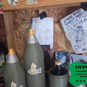 May include: Three large, olive-green cylindrical objects with orange and silver tops, adorned with stickers. A bottle with a wooden stopper sits nearby. Handwritten notes and drawings are visible on the wooden background.