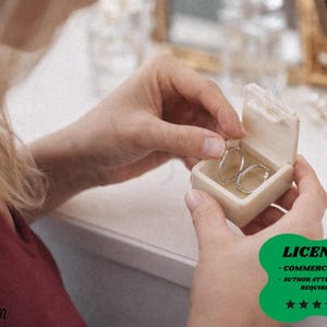 Könnte beinhalten: Eine kleine, cremefarbene Schmuckschatulle wird geöffnet, um ein Paar silberner Creolen zu enthüllen. Die Ohrringe sind mit kleinen, funkelnden Steinen besetzt. Die Box wird von einer Person gehalten, die ein rotes Oberteil trägt.