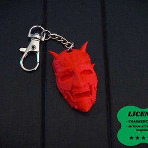 May include: A red devil face keychain with a silver clasp and chain. The devil face has horns and a detailed, textured surface. The keychain is on a dark background. A green sticker with the word "LICENSE" is in the bottom right corner.