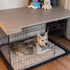 Peut inclure: Une cage pour chien en fil de fer noir avec un dessus en bois gris clair. Un chien au pelage beige et gris est à l'intérieur de la cage, couché sur un lit beige. Une plante en pot et des peluches sont visibles en arrière-plan.