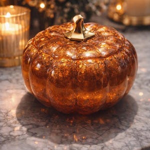 Può includere: Una decorazione a forma di zucca in vetro color ambra, caratterizzata da una finitura interna testurizzata e scintillante e da un gambo liscio color oro metallizzato. La zucca presenta una superficie a coste e un aspetto traslucido.